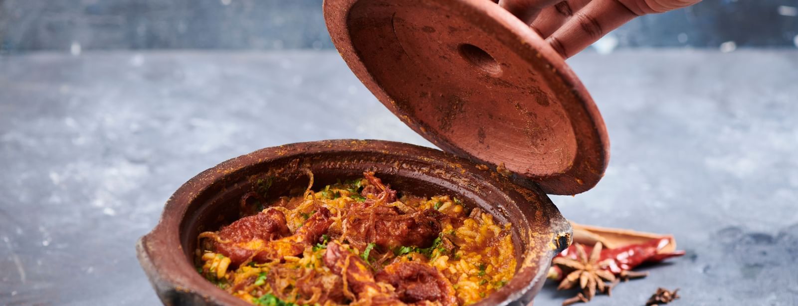 A hand opening the lid of a clay pot filled with biryani at Grande Bay Resort & Spa.