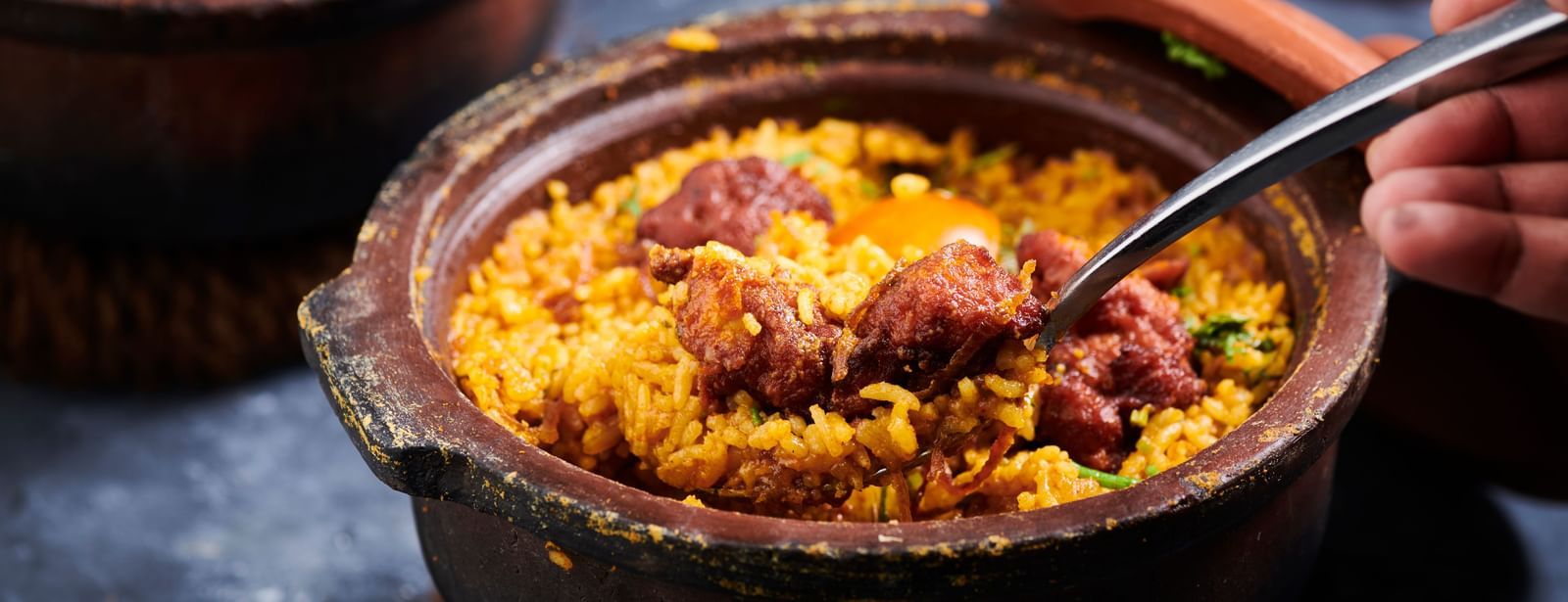 A hand scooping biryani from a clay pot with a spoon at Grande Bay Resort & Spa.