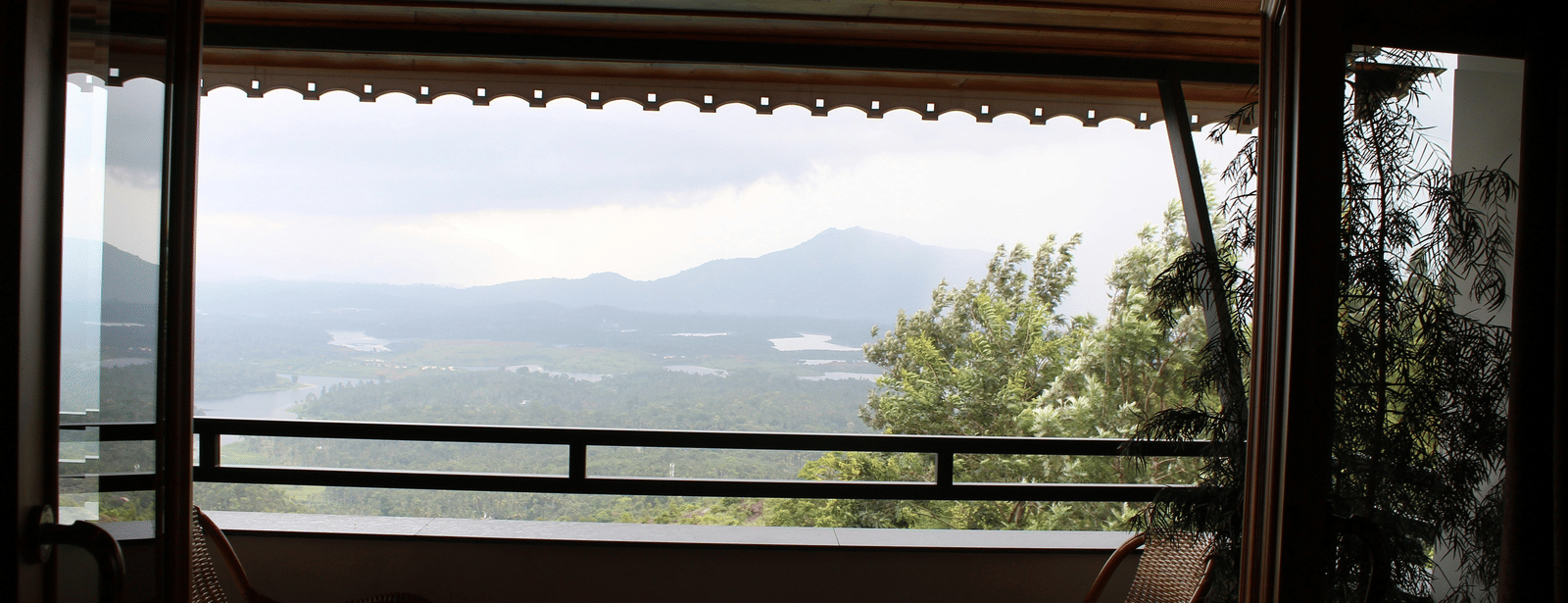 A wide-angle view from a dark indoor area onto a balcony with wicker chairs, overlooking a vast, lush green valley.