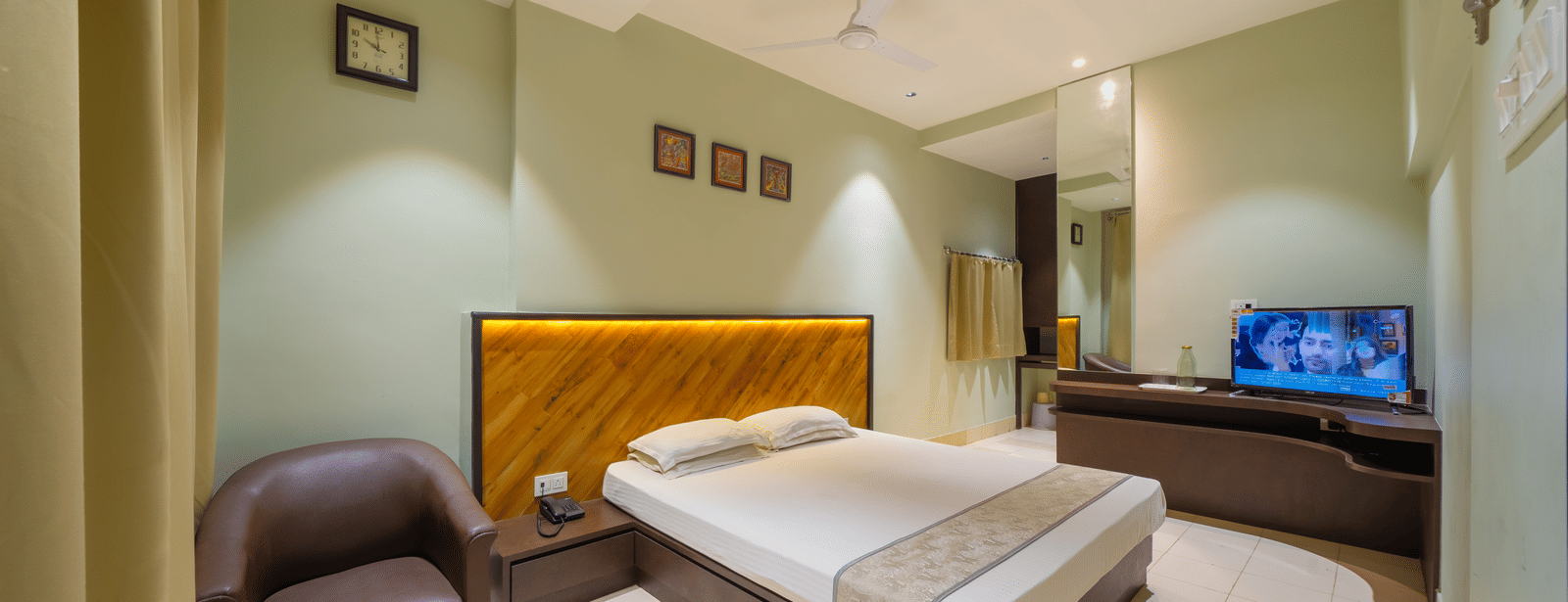 A spacious hotel bedroom featuring a double bed with a headboard, light green walls, a TV, and a leather armchair - Hotel Dolphin Puri