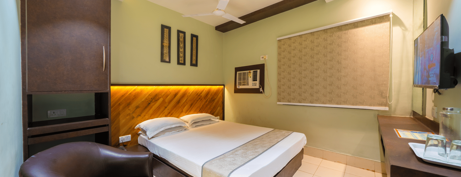 A hotel bedroom with light green walls, a double bed, a dark wood headboard, a round leather armchair, and a small desk - Hotel Dolphin Puri