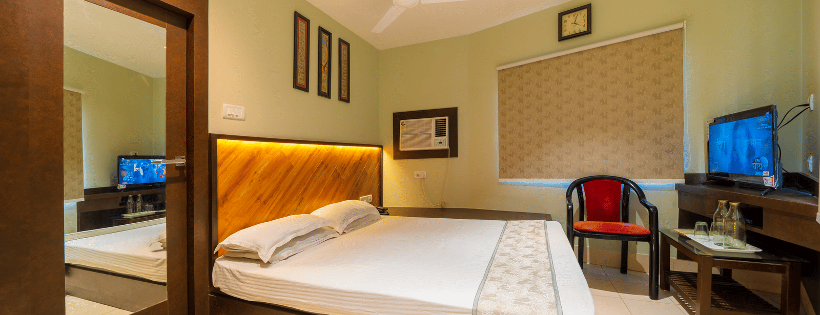 A spacious hotel bedroom showing the double bed, a wooden wardrobe with a full-length mirror, a closed blind on the window, and a TV - Hotel Dolphin Puri