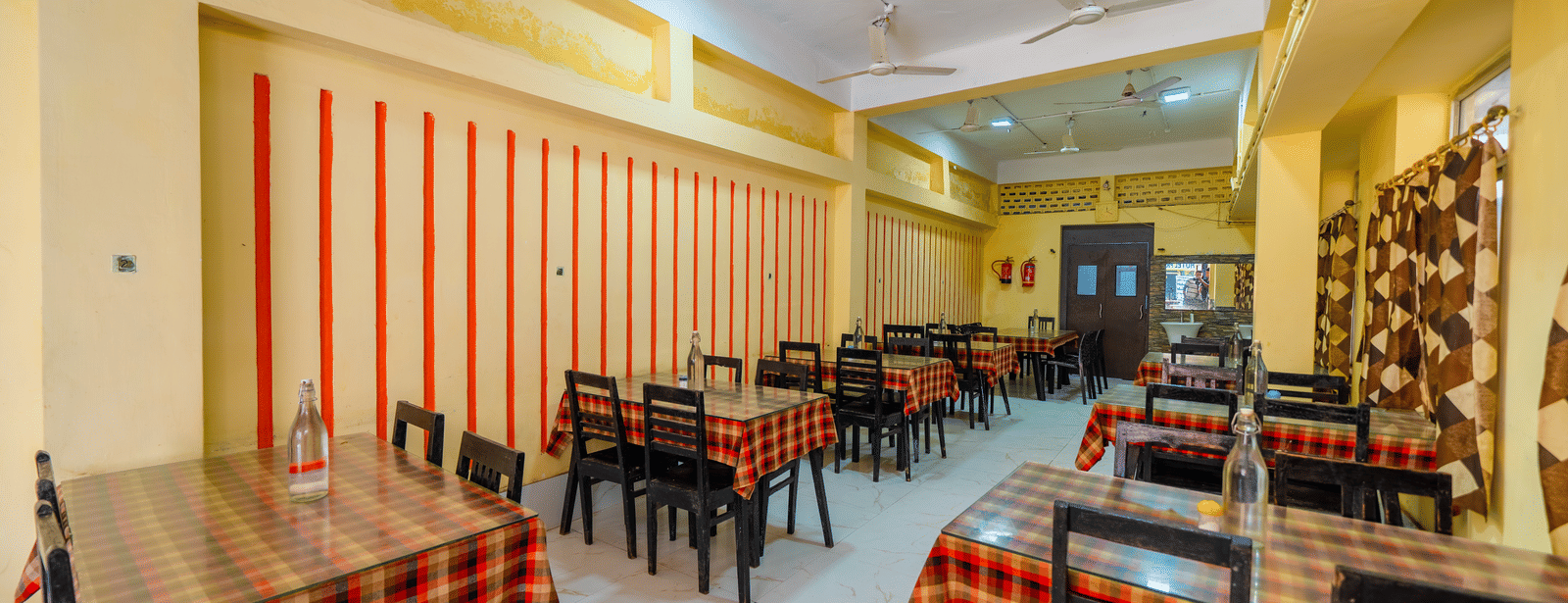 A brightly-lit, empty hotel dining hall with small tables covered in red and black checkered tablecloths and matching chairs - Hotel Dolphin Puri