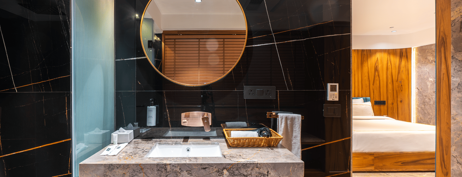 A washbasin with circular mirror, black tiled wall and toiletries at Infiniti Hotel, Indore.