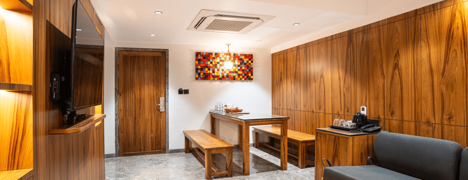 A suite with wooden walls, grey sofa, dining table, television and decorative painting at Infiniti Hotel, Indore.