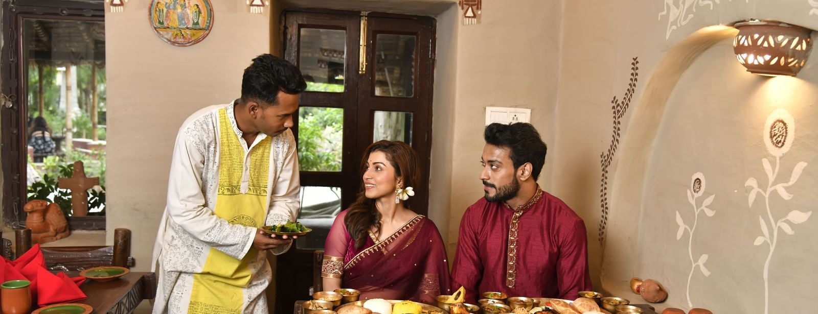 A server in traditional attire presenting food to two seated individuals at a low table | Bhoomi Restaurant