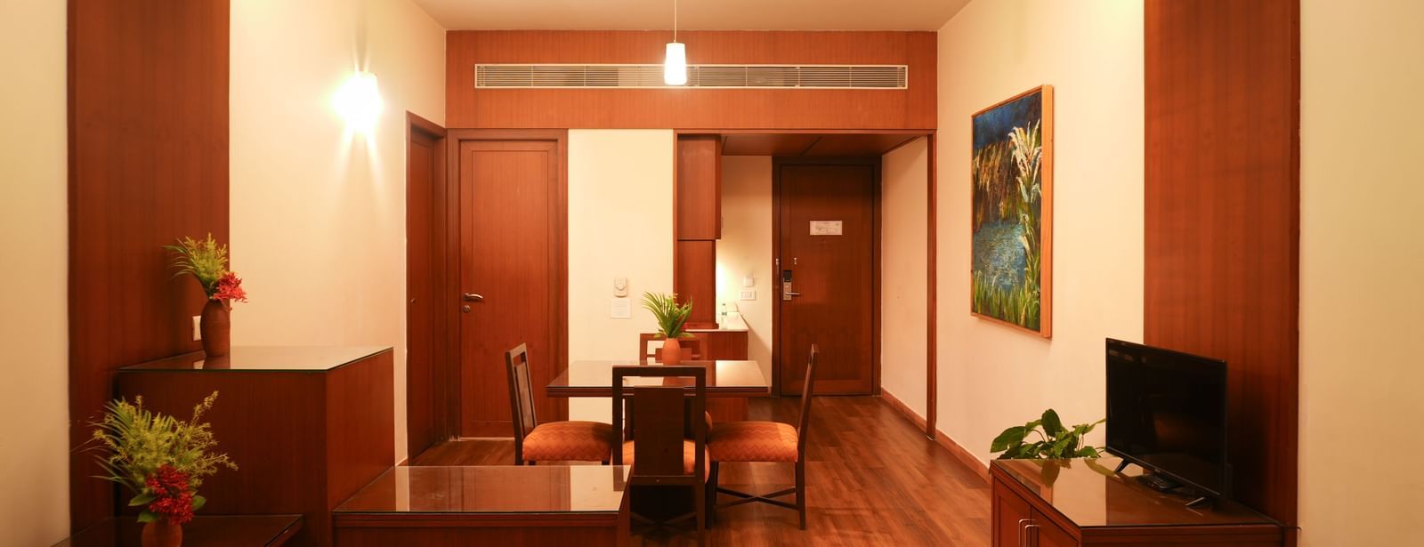 Dining and entertainment space in the One-Bedroom Suite at Vedic Village Spa Resort with ambient lighting, wooden furnishings, flat-screen TV on a table, potted plants, and framed wall art.