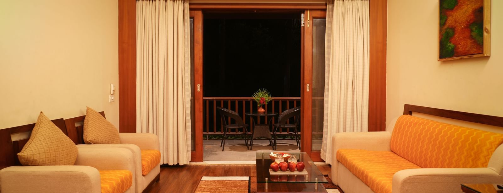 Plush sofas in the living room of the One-Bedroom Suite at Vedic Village Spa Resort, set against carpeted wooden floors and opening to a balcony with a wooden centre table.