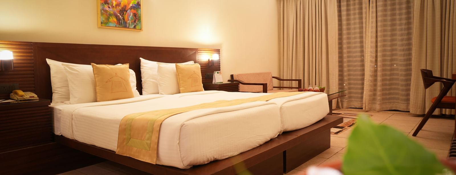 Neatly arranged double bed in the Standard Suite at Vedic Village Spa Resort with a wooden headrest, a painting above the bed, and large window drapes drawn shut.