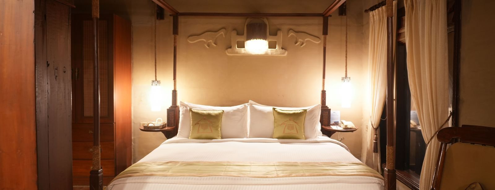 Elegantly styled four-poster king size bed in the Zamindar Suite at Vedic Village Spa Resort, complemented by a wooden lounge chair, and soft lighting.