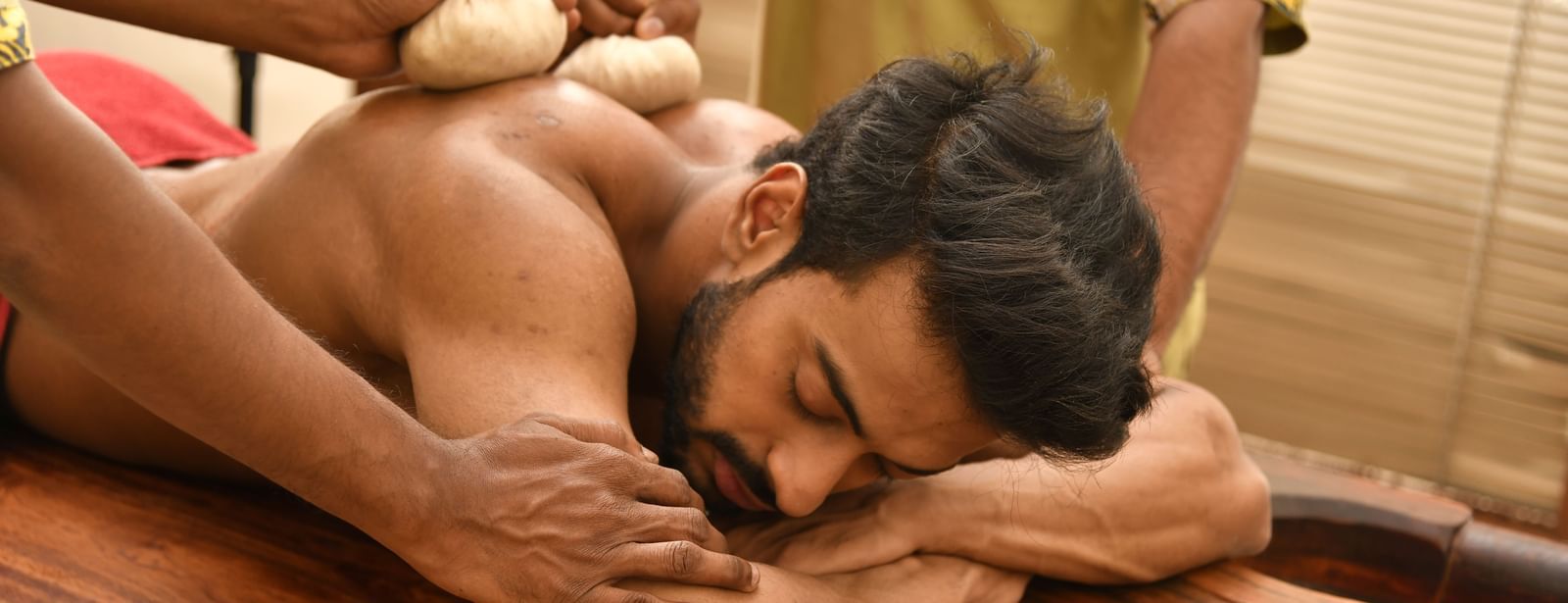 A man receiving a traditional Ayurvedic massage with herbal pouches, lying face down on a wooden massage table