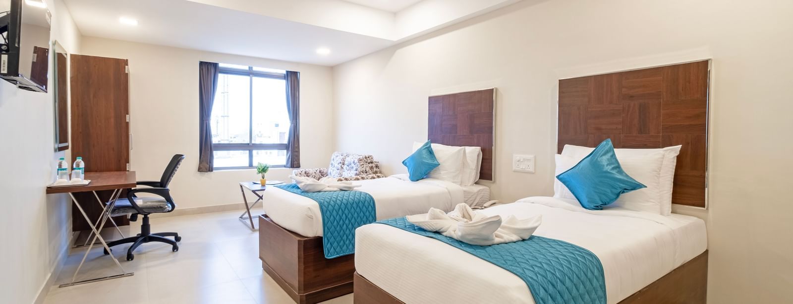 Twin beds with fresh linens blue runners seating area and bright ambience creating comfort at Swarna's The Capital.