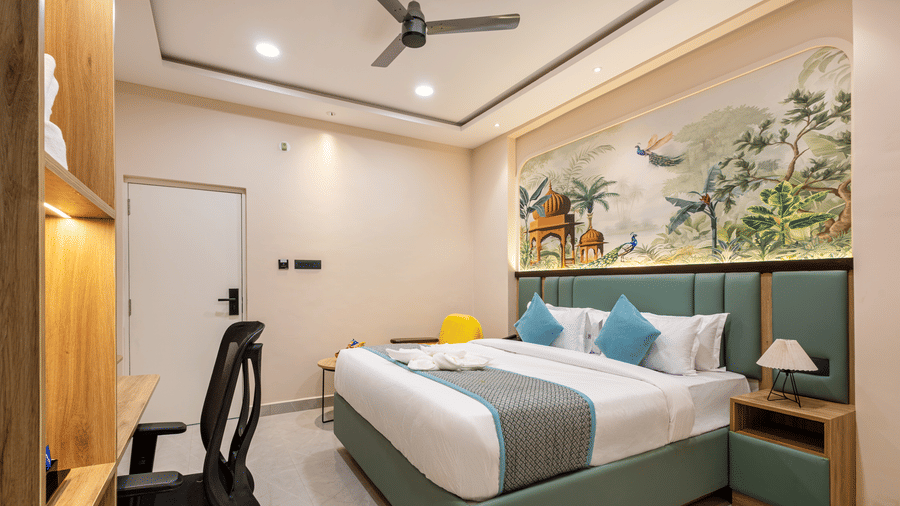 A bedroom setup displaying a king size bed with blue pillows, a desk area with a chair, and a wall mural at Monday Hotels & Suites Madhapur Hyderabad.