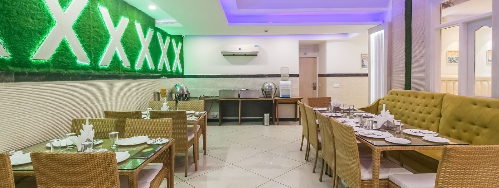 A restaurant interior with a long tufted sofa, wooden tables and chairs, a decorative wall design, and a tiled floor, lit by soft lighting. - Hotel Diamond Plaza, Chandigarh.
