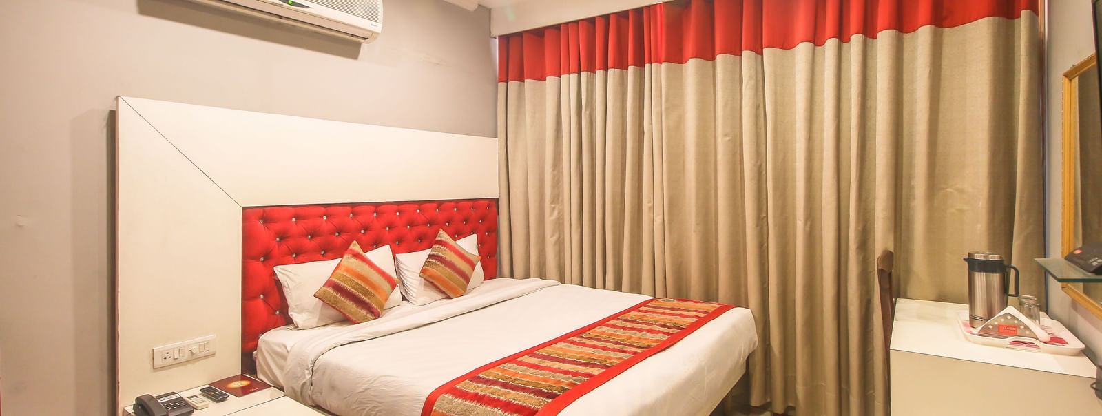 A hotel room with a plush bed featuring a patterned runner, layered curtains, a white headboard, and an AC unit, illuminated by soft lighting. - Hotel Diamond Plaza, Chandigarh.