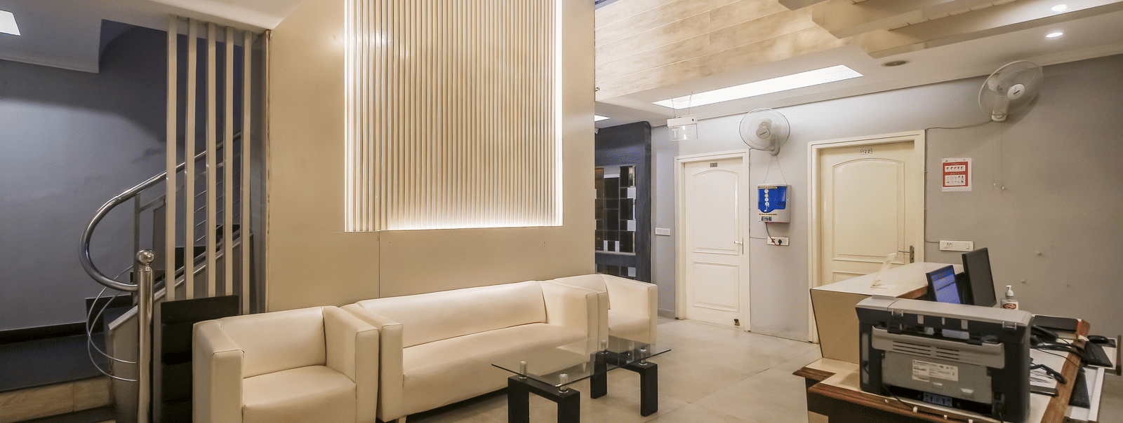 A hotel lobby with a seating area featuring white sofas, a textured wall with lighting, a spiral staircase, and a reception desk with equipment. - Hotel Diamond Plaza, Chandigarh.