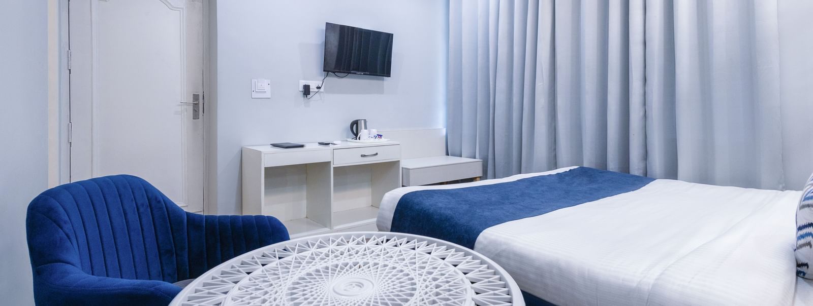 Stylish hotel room with a bed, white ornate circular table, blue accent chairs, and grey curtains at Hotel Downtown17, Chandigarh.