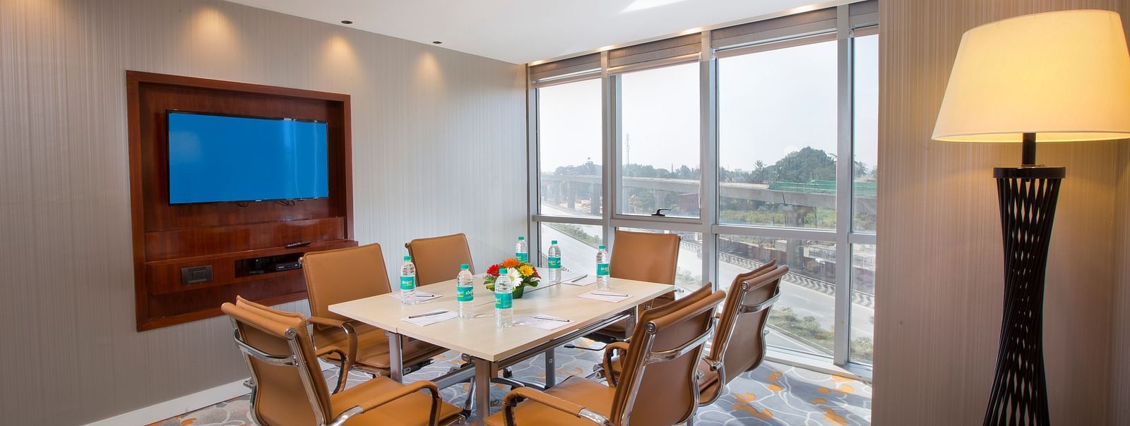 A small meeting room with a round table, tan leather chairs and a TV, with a large window and a floor lamp - Hotel Southend by TGI - Bommasandra, Bangalore