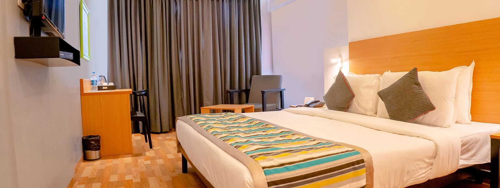Another view of a cosy hotel room with a large bed, colourful throw and wooden furniture, including a desk with a TV above it - TGI Ameya Grand, Goa
