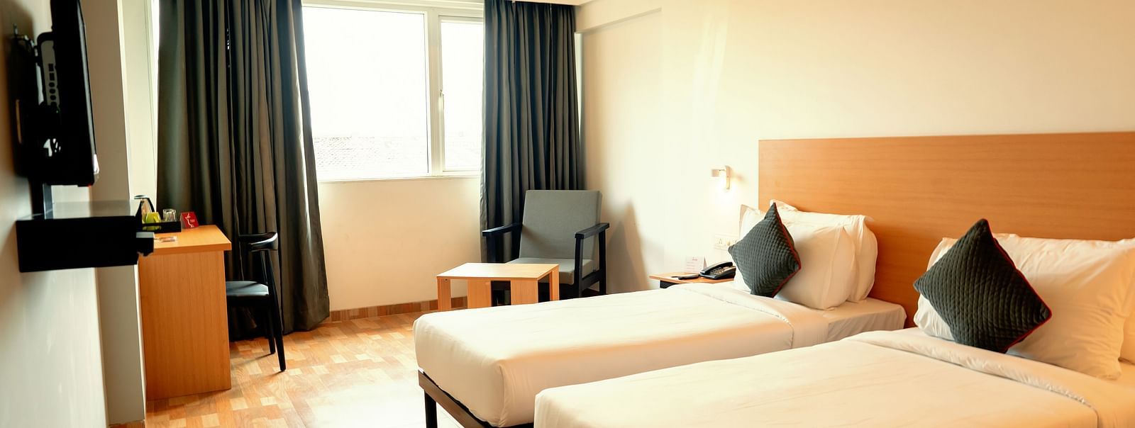 A bright hotel room with two single beds, a desk and a flat-screen TV, with a large window draped in gray curtains - TGI Ameya Grand, Goa