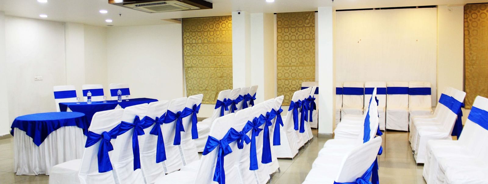 A banquet hall with chairs tied with blue ribbons and round tables with blue runners - TGI Apple Inn, Jaipur 