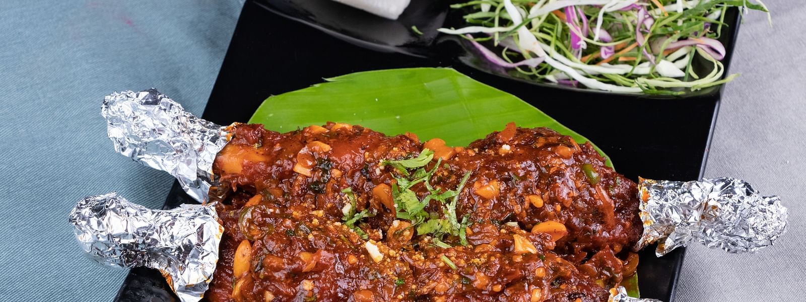 Grilled meat entree served on a leaf-shaped plate, decorated with herbs and foil-wrapped bones. - The President Hotel, Kumara Park, Bangalore