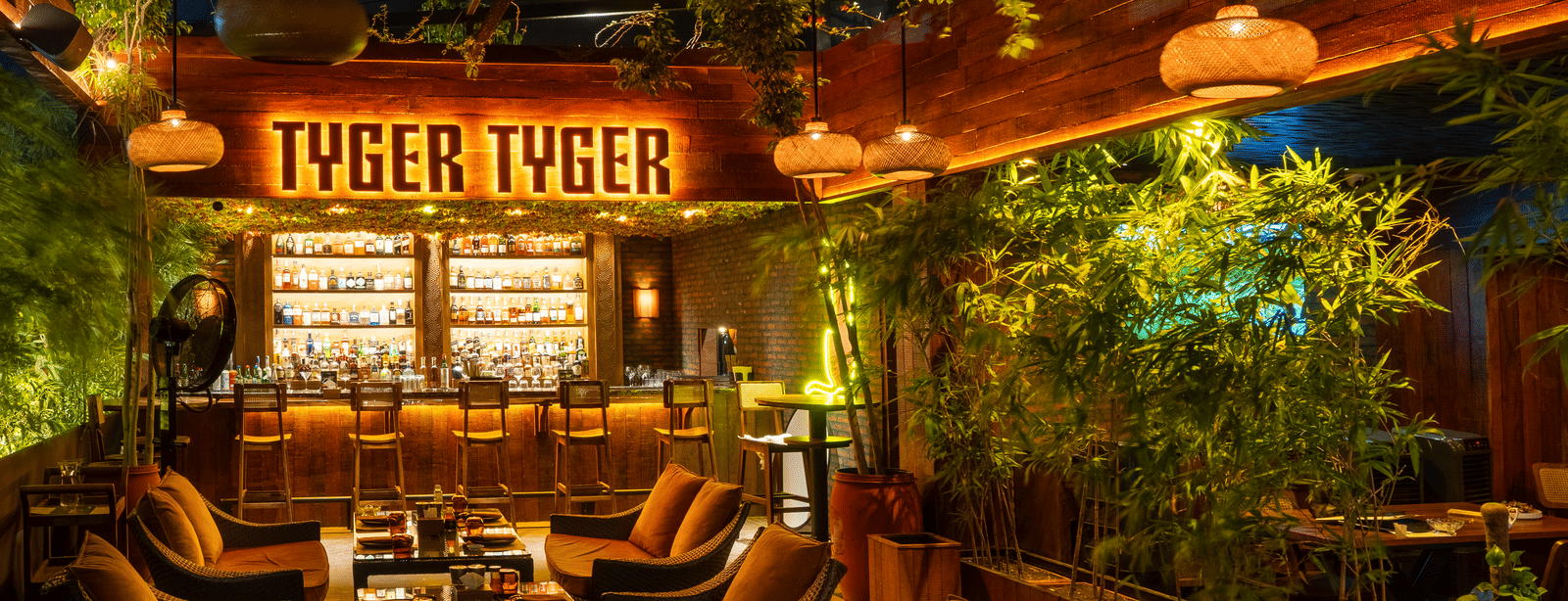A restaurant seating area in a greenery setting with brown sofas, hanging lights and a signboard reading 'TYGER TYGER' at Infiniti Hotel, Indore.