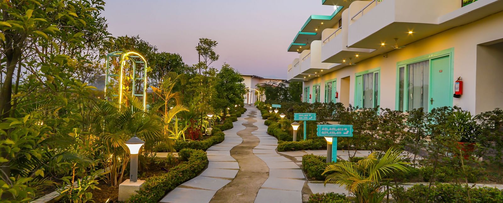 A pathway illuminated at dusk winds through the grounds of Vedikant Resorts The Mallard Corbett, showcasing the buildings.