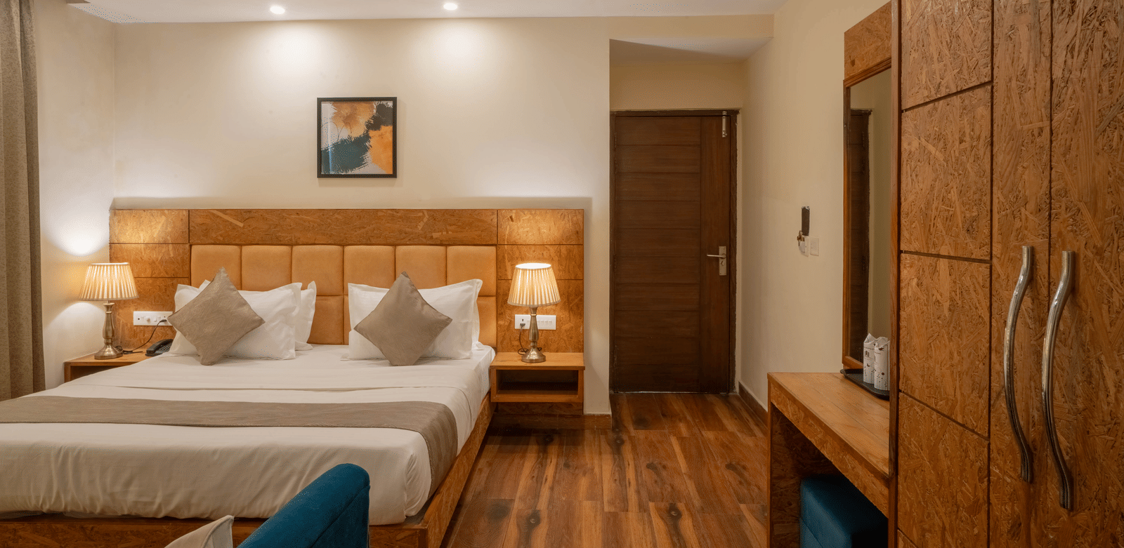 A hotel room, featuring a large bed with a beige headboard, a dark wooden wardrobe, a mounted mirror, and a small blue chair | Perfectstayz Value Valley Vista
