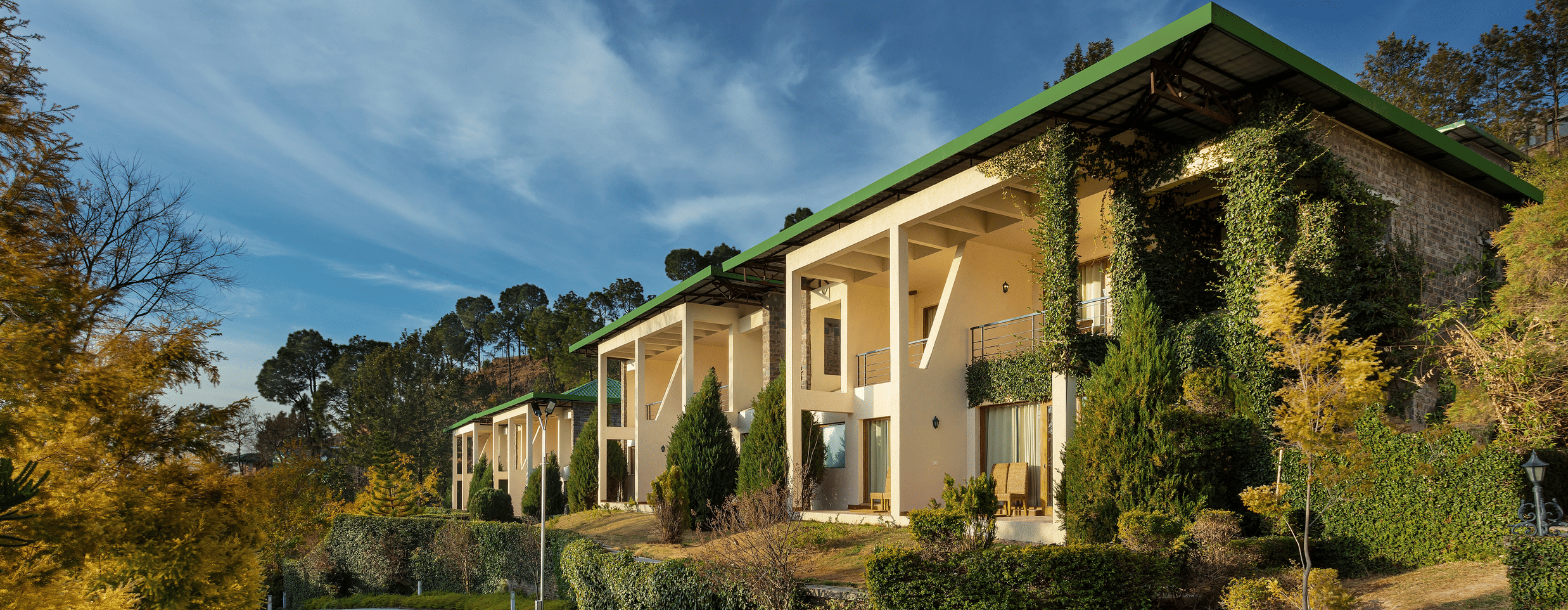 Facade of Suryavilas Luxury Resort and Spa in Solan with a paved driveway and green climbing plants on its exterior walls, under a blue sky with clouds