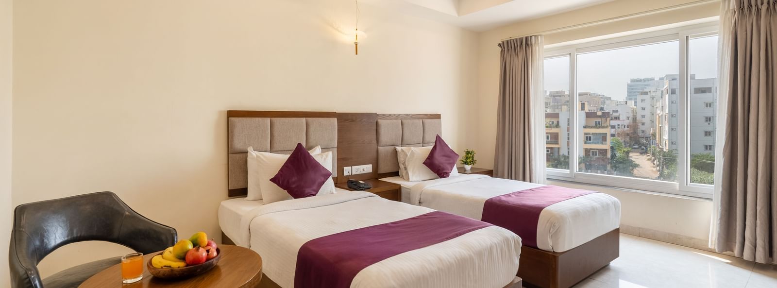 A Deluxe Room at Destino by Monday Hotels, Gachibowli, with twin beds, a small table with a fruit basket, and a modern chair.