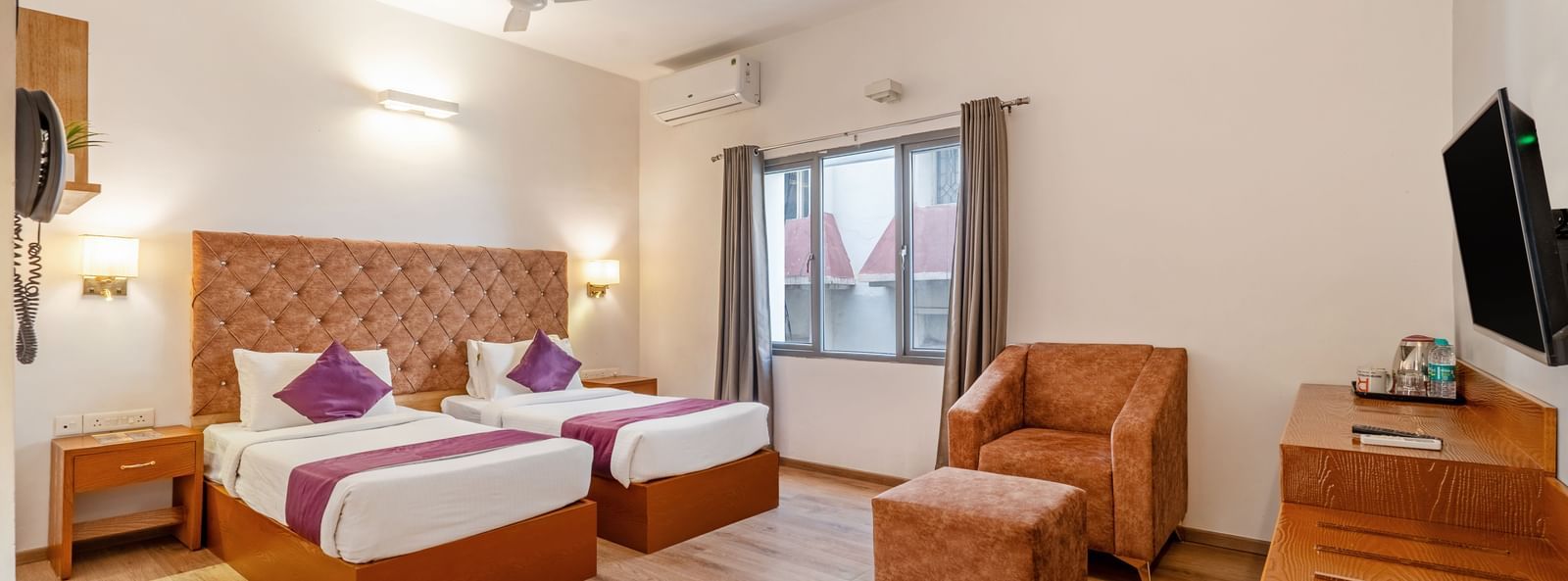 A bright hotel room with 2 single beds, cushioned headboards, a TV, a wooden desk, chairs, and a large window – Monday Hotels HITEC City, Hyderabad