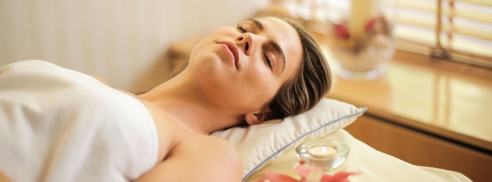 A person lies relaxed on a spa bed, eyes closed, with a towel draped over them and flower petals nearby