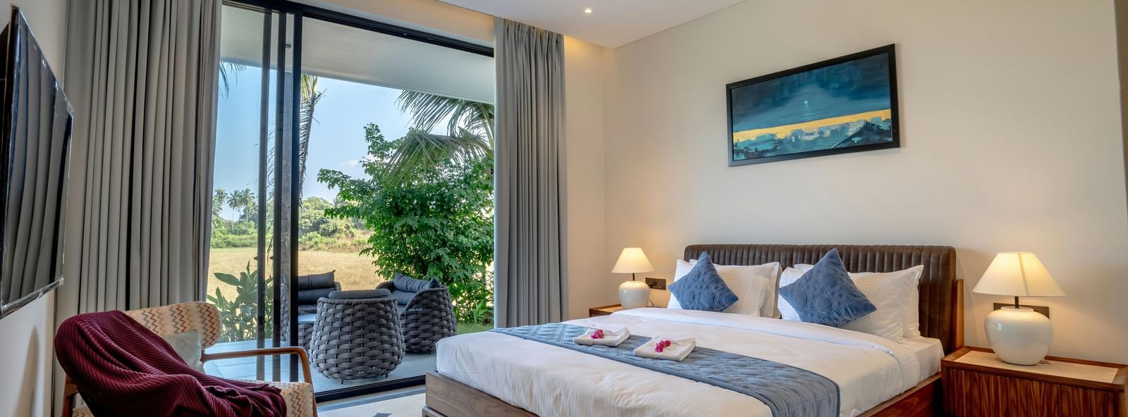 A room with balcony, spacious bed with bedside tables and lamps, artwork above the headboard, and outdoor view through glass doors
