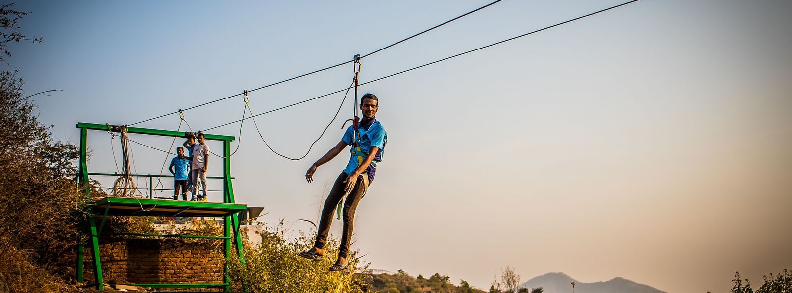 A person zip-lining across a scenic landscape with hills in the background.- Bamboo Saa Resort & Spa, Udaipur