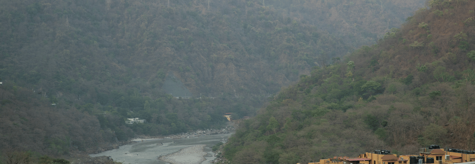 A panoramic view of a valley at Perfectstayz Premium Laxmi Heritage, featuring a wide riverbed, forested hillsides, and a building in the foreground.