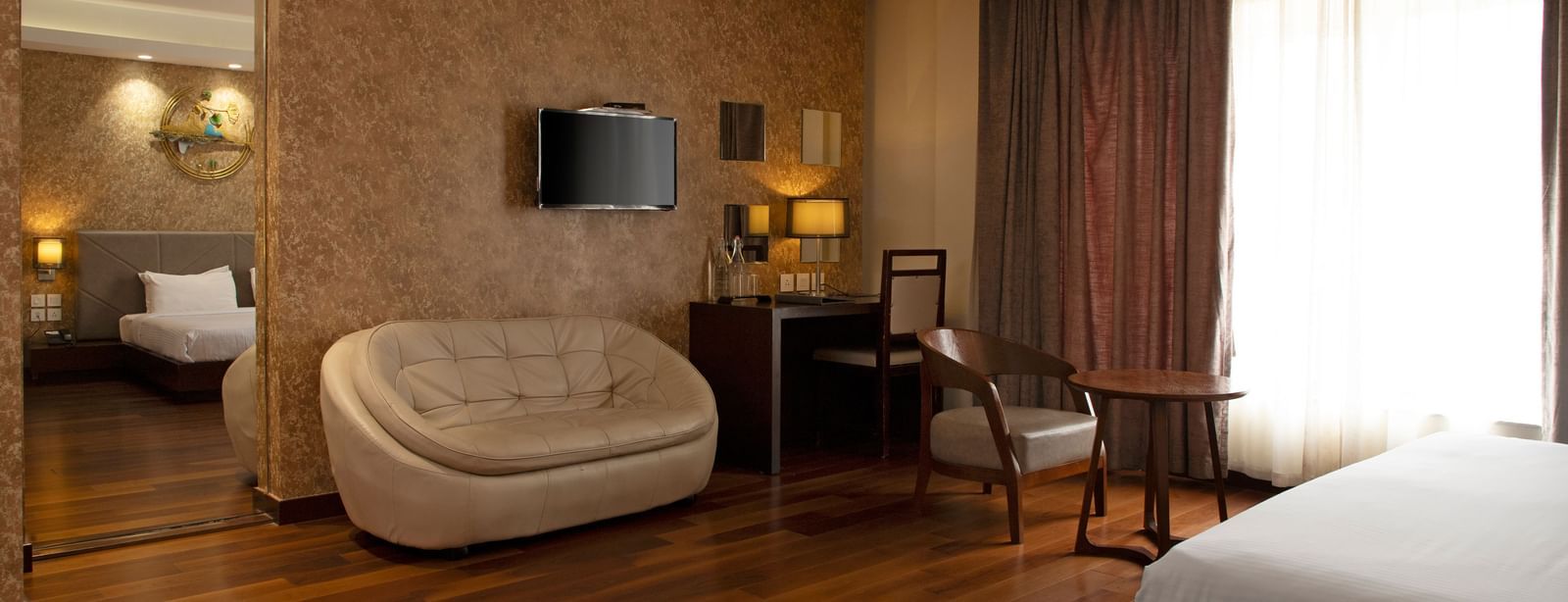 A spacious room featuring a large bed, a sofa, a flat-screen TV mounted on the wall, and wooden flooring at The Hamuse Hotel, Kodaikanal.