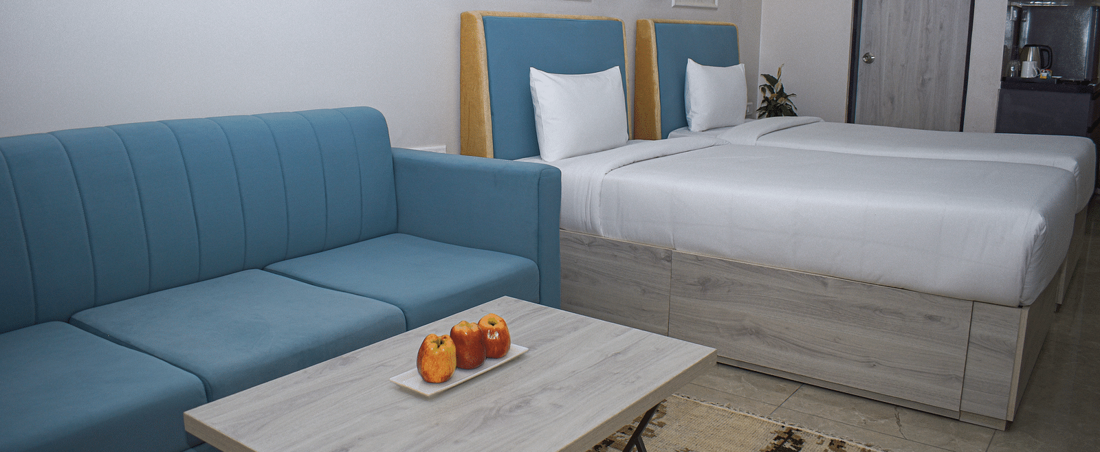 A hotel room with two twin beds, a blue sofa, a light wood coffee table with fruit, and a kitchenette area - Urban Suites by BluSalzz, Jaipur