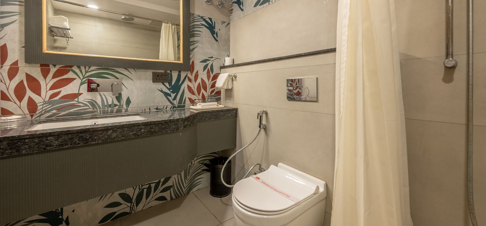 A bathroom with floral wallpaper, washbasin, mirror, and a toilet next to a shower with curtain at Hotel Highway King, Jaipur.