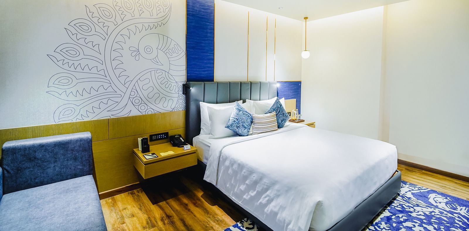 An image of our Deluxe Suite in Madurai with a peacock wall paper, a large bed, a night stand and a blue couch