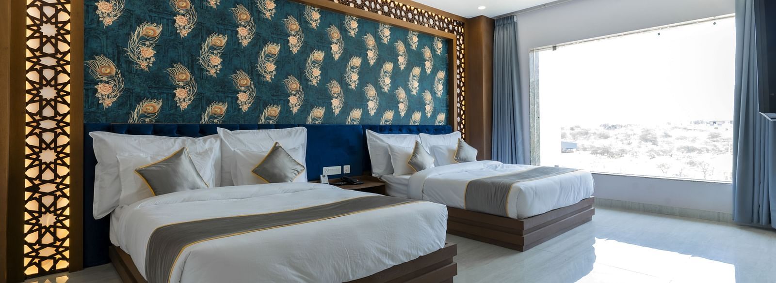 A bedroom at the Bamboo Saa Mulberry Resort featuring two twin beds with white linens and grey throws. A decorative wall panel with a blue and gold pattern is above the beds and a large windows is visible.