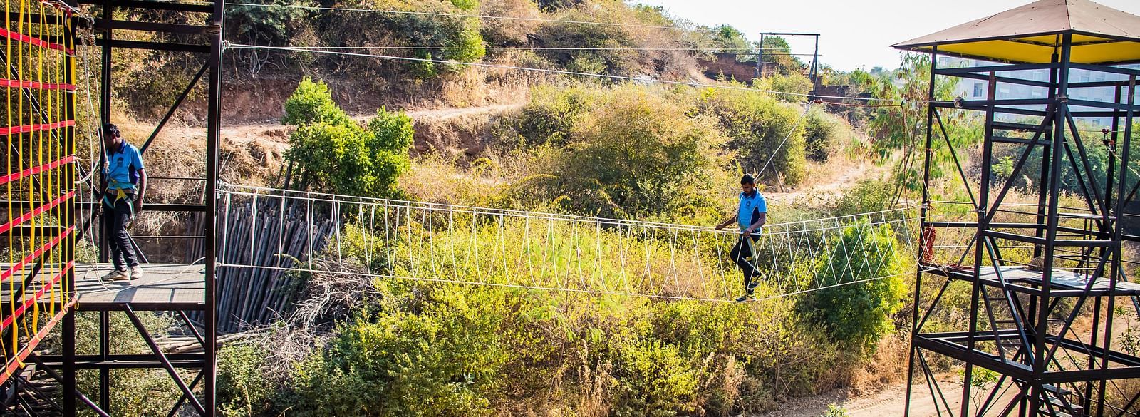 Two people zip-lining across a scenic landscape with hills and trees.- Bamboo Saa Resort & Spa, Udaipur