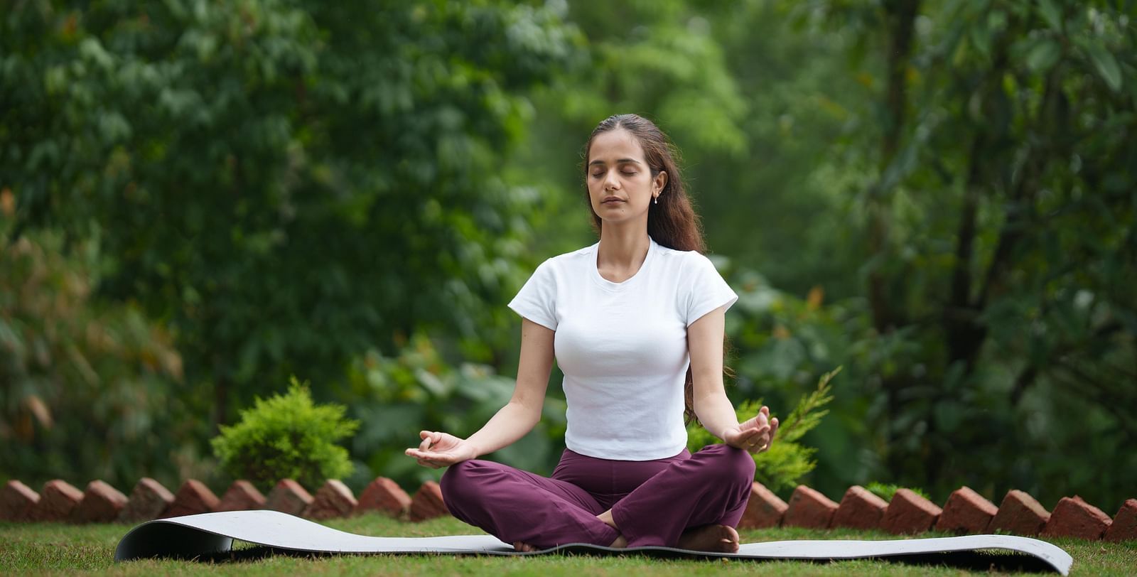 A woman in a white t-shirt and purple yoga pants sits in a meditative pose on a yoga mat outdoors amidst lush green trees, likely at  Summit Enigma Resort and Spa.