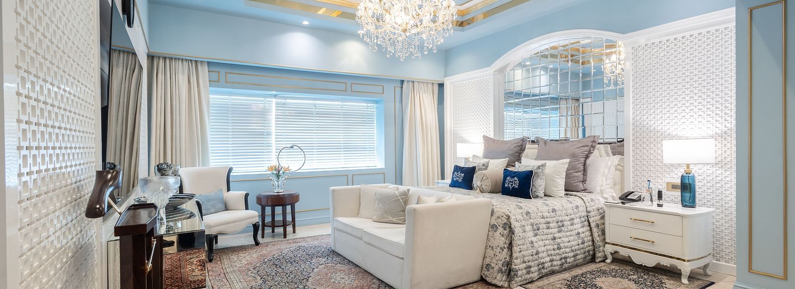 An image of a suite with a large bed and a sofa placed in front of a television and a chandelier hanging from the ceiling