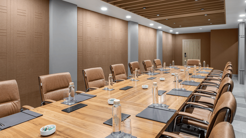 A conference room featuring a long, light wood meeting table surrounded by brown upholstered chairs, with wood-panelled walls and a matching suspended ceiling section | Voyage Eastin Suites