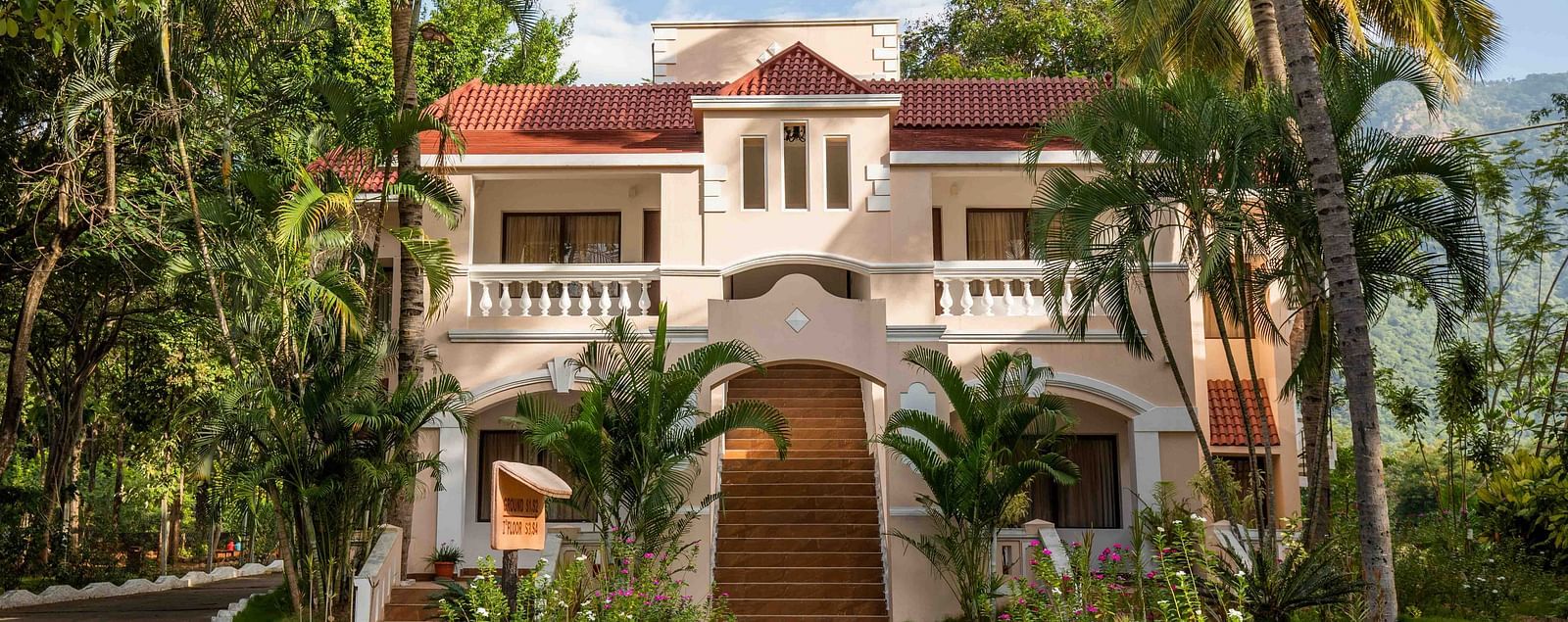 A grand two-storey villa-style building at Avana Resort with a terracotta roof, surrounded by palm trees and lush gardens under a bright sky.