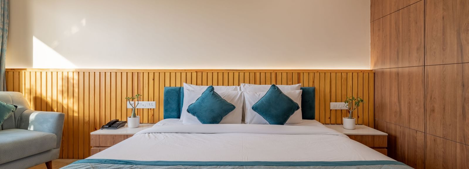 Hotel Highway King, Bagru, bedroom with bedside tables, cushions, armchair, and wooden paneled wall.