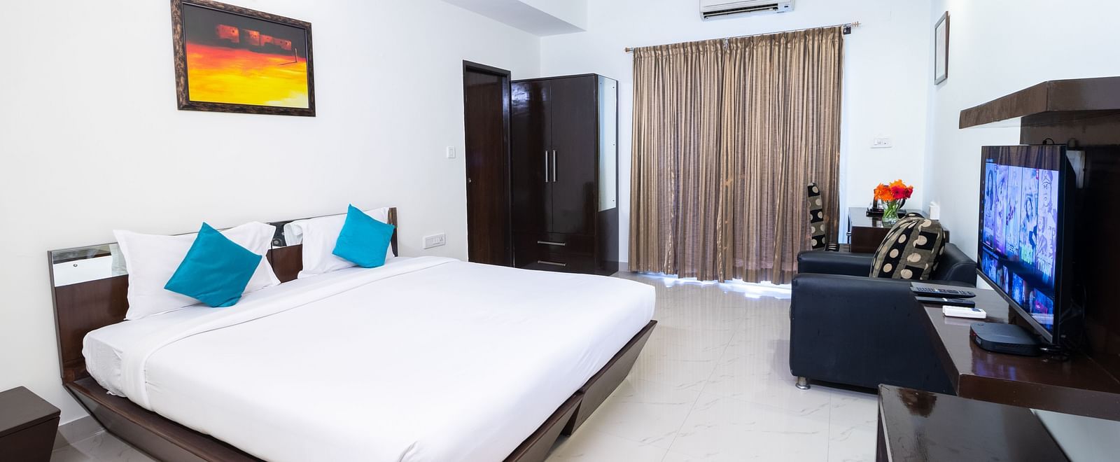A room offering a double bed, a wall-mounted television, a sofa, and an artwork hung on the wall at Sanctum Suites Domlur, Bangalore.