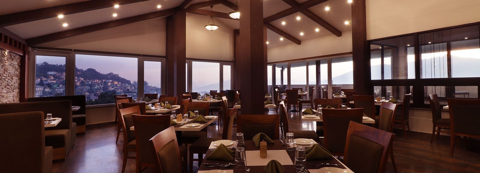 A modern styled rooftop dining space featuring wooden furniture and arranged table set with tableware and folded napkins and huge windows that opens to hilly cityscape at the backdrop.