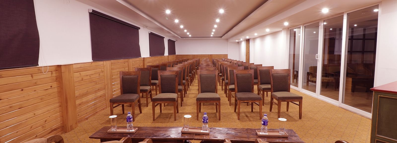 A view of a panel with three chairs and two table featuring water bottles and a pamphlet overlooking the multiple chairs arranged in rows at the backdrop.